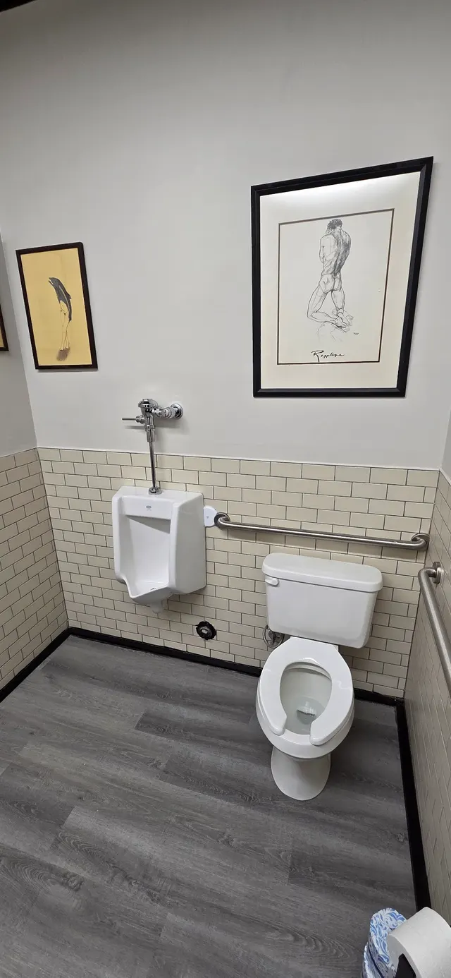 A commercial restroom with a wall-hung urinal, a toilet with grab bars, white subway tile and grey plank flooring.