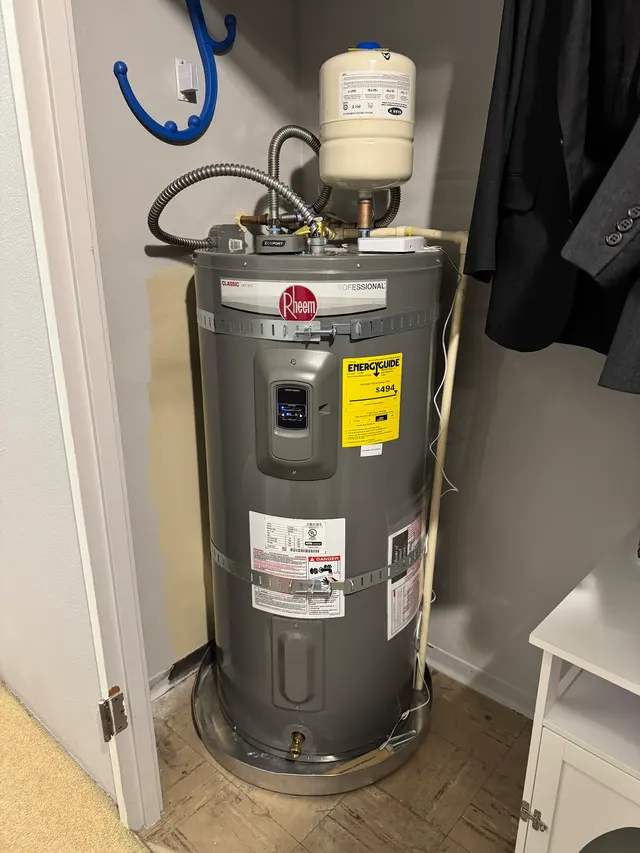 A new Rheem Professional electric water heater in a closet, with an expansion tank, flexible connectors and the EnergyGuide label still attached.