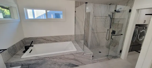 A finished bathroom with a marble-clad soaking tub, matte black tub filler and a glass-enclosed marble shower, with a laundry room beyond.