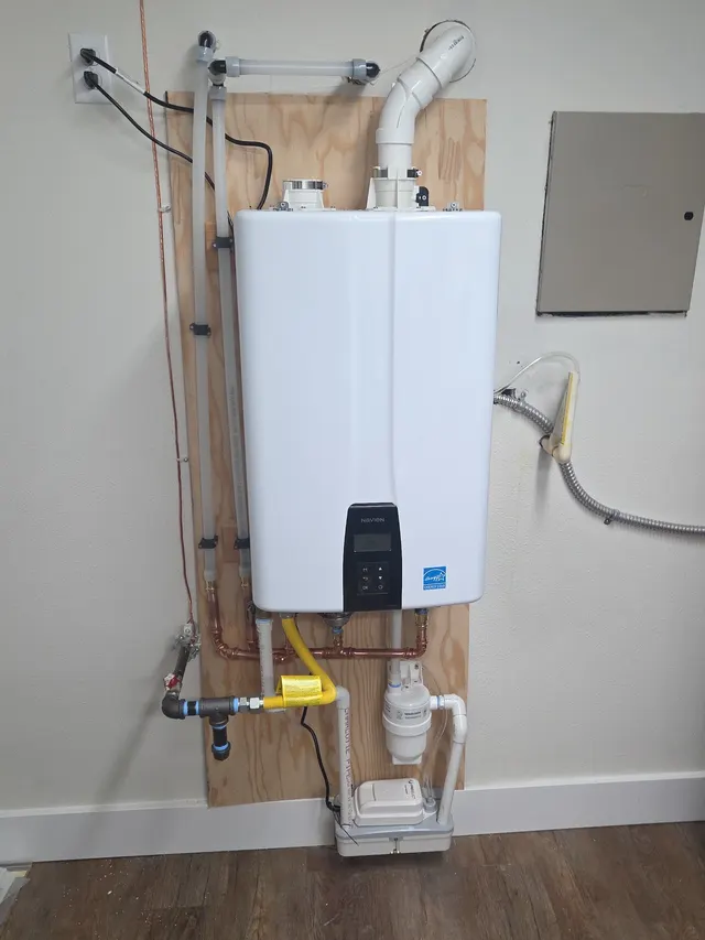 A wall-hung Navien condensing tankless water heater installed on a plywood backer, with new copper supply lines, a yellow gas flex connector, PVC venting and a condensate pump below.