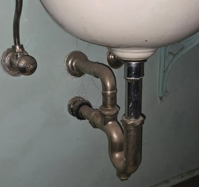 The corroded chrome P-trap and supply valve under an old wall-hung sink.