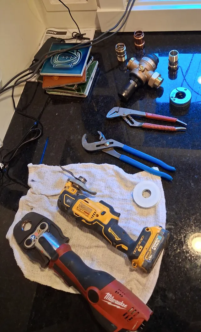 A new brass pressure-reducing valve and press fittings laid out on a counter with a Milwaukee press tool, a DeWalt multi-tool, two pairs of pliers and a roll of thread tape, ready to install.