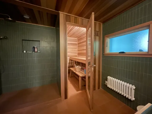 A green-tiled walk-in shower beside a cedar sauna, with a radiator under a frosted window.